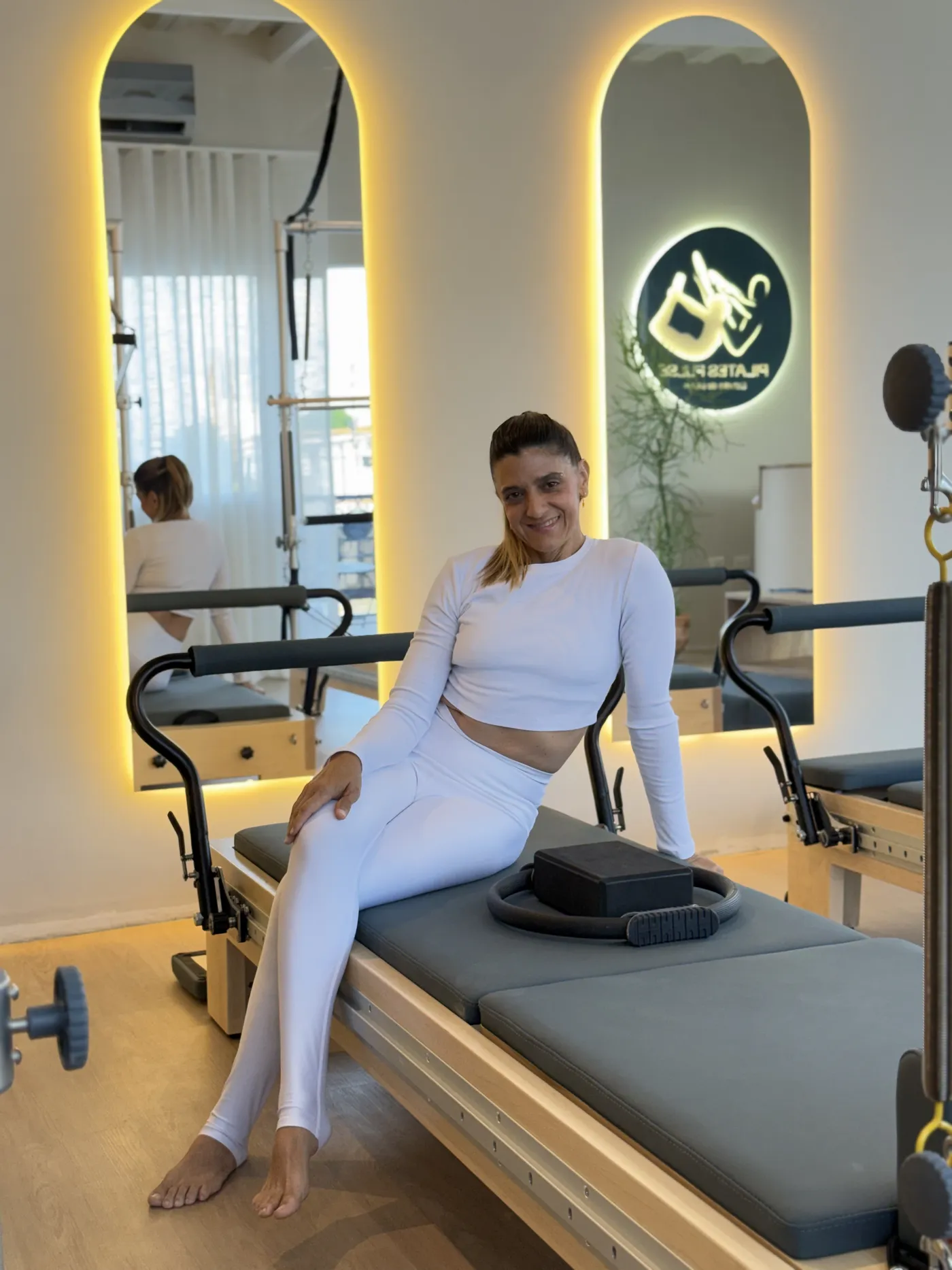 La instructora principal sentada en un reformer, con el logo de Pilates Pulse iluminado en la pared de fondo